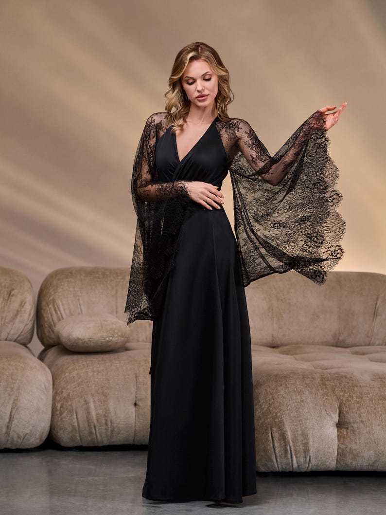 May include: A black, floor-length dress with a V-neck and flowing skirt. The dress features sheer, black lace sleeves that extend into a cape-like design. The model is standing in front of a neutral-toned background.