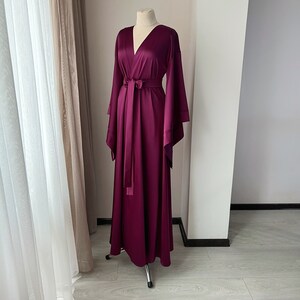 May include: A long, burgundy satin robe with a V-neckline and wide, flowing sleeves. The robe has a sash that ties at the waist.