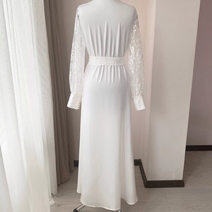 Puede incluir: Un vestido blanco largo hasta el suelo con mangas largas de encaje transparente con motivos florales y pu&ntilde;os abotonados. La prenda tiene cuello alto, cintura entallada con una banda ancha y una falda lisa y opaca que cae recta.