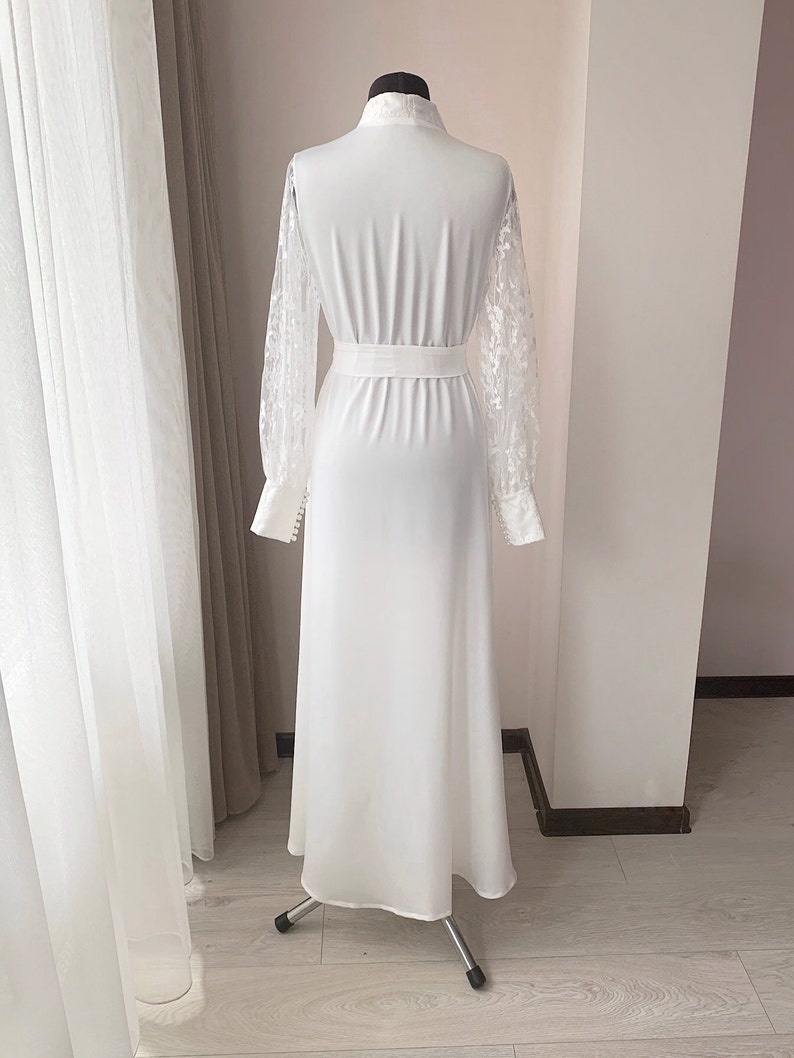 Puede incluir: Un vestido blanco largo hasta el suelo con mangas largas de encaje transparente con motivos florales y pu&ntilde;os abotonados. La prenda tiene cuello alto, cintura entallada con una banda ancha y una falda lisa y opaca que cae recta.