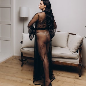 May include: A woman wearing a black sheer robe with a lace trim. The robe is open in the front, revealing a nude lace bra and matching underwear. The woman is standing in a room with a white couch and a lamp in the background.