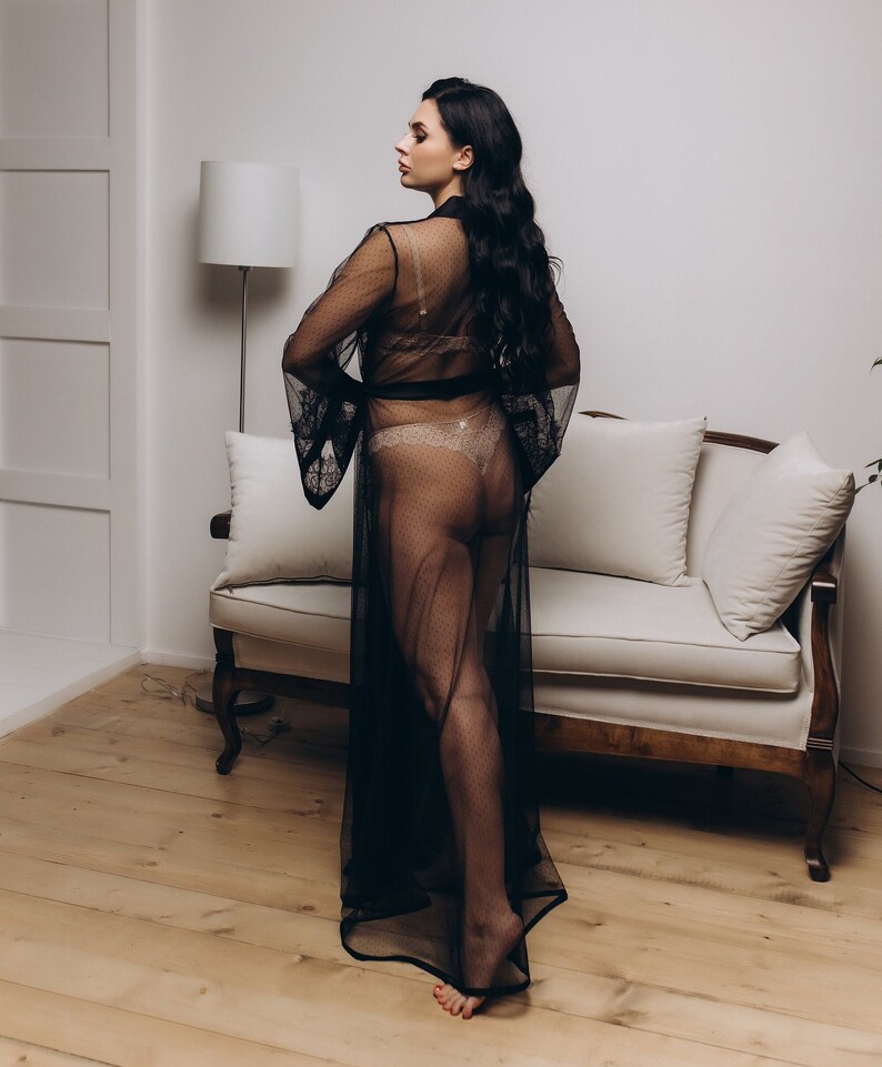 May include: A woman wearing a black sheer robe with a lace trim. The robe is open in the front, revealing a nude lace bra and matching underwear. The woman is standing in a room with a white couch and a lamp in the background.