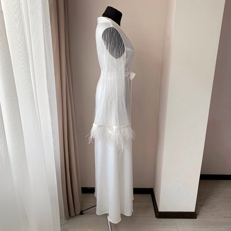 May include: A floor-length white robe featuring long, sheer sleeves with delicate fringe detailing and a matching white tie belt at the waist. The garment is displayed on a mannequin, showcasing a smooth, solid fabric construction with feather-like trim.