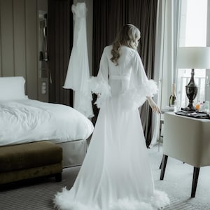 May include: A floor-length white robe featuring long, wide sleeves and a hemline trimmed with white feathers. The robe has a wrap-style front with a matching tie belt at the waist, creating a flowing silhouette that reaches the floor.