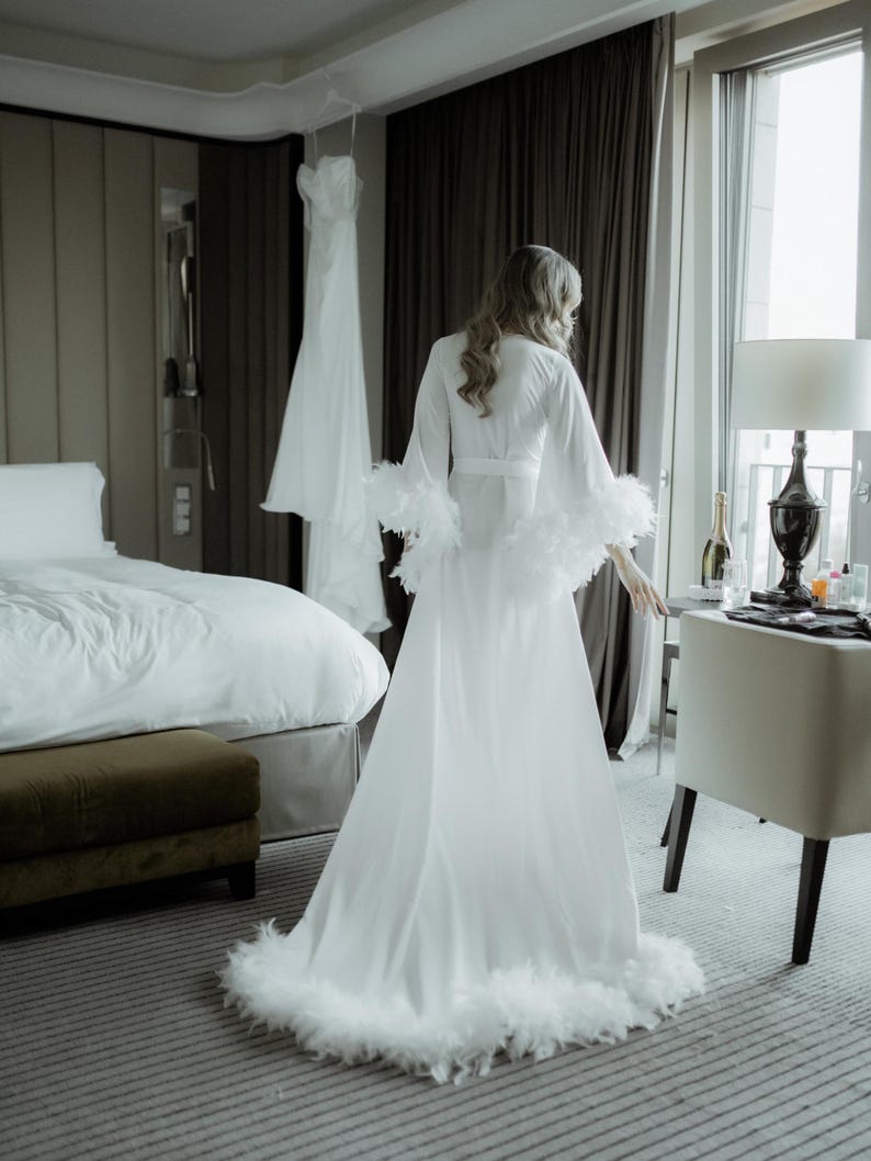 May include: A floor-length white robe featuring long, wide sleeves and a hemline trimmed with white feathers. The robe has a wrap-style front with a matching tie belt at the waist, creating a flowing silhouette that reaches the floor.