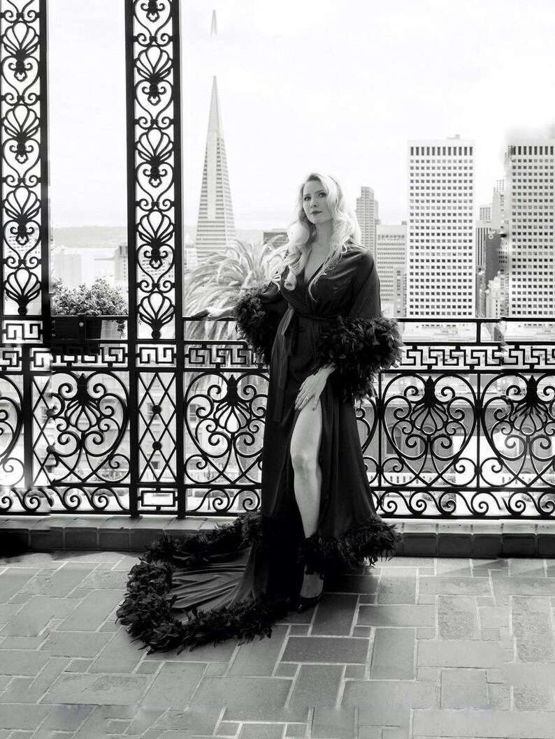 May include: A woman wearing a black robe with feather trim. The robe is open in the front and has a long train. The woman is standing on a balcony with a city view in the background.
