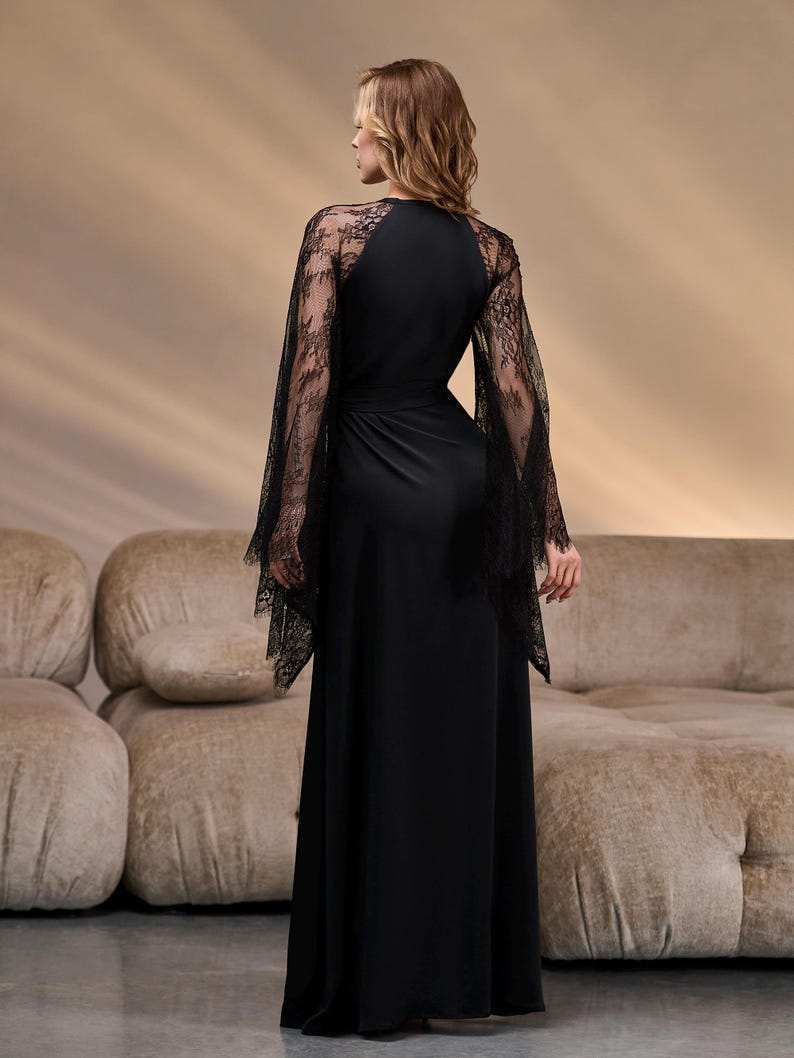 May include: A long black dress with lace sleeves. The dress has a fitted bodice and a flowing skirt. The sleeves are made of delicate black lace. The model is standing in front of a beige sofa.