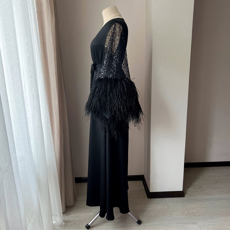 K&ouml;nnte beinhalten: Ein schwarzes Maxikleid mit langen &Auml;rmeln, die mit Pailletten besetzt sind und einen transparenten &Uuml;berzug aus Chiffon sowie einen Federnbesatz an den &Auml;rmelb&uuml;ndchen haben.