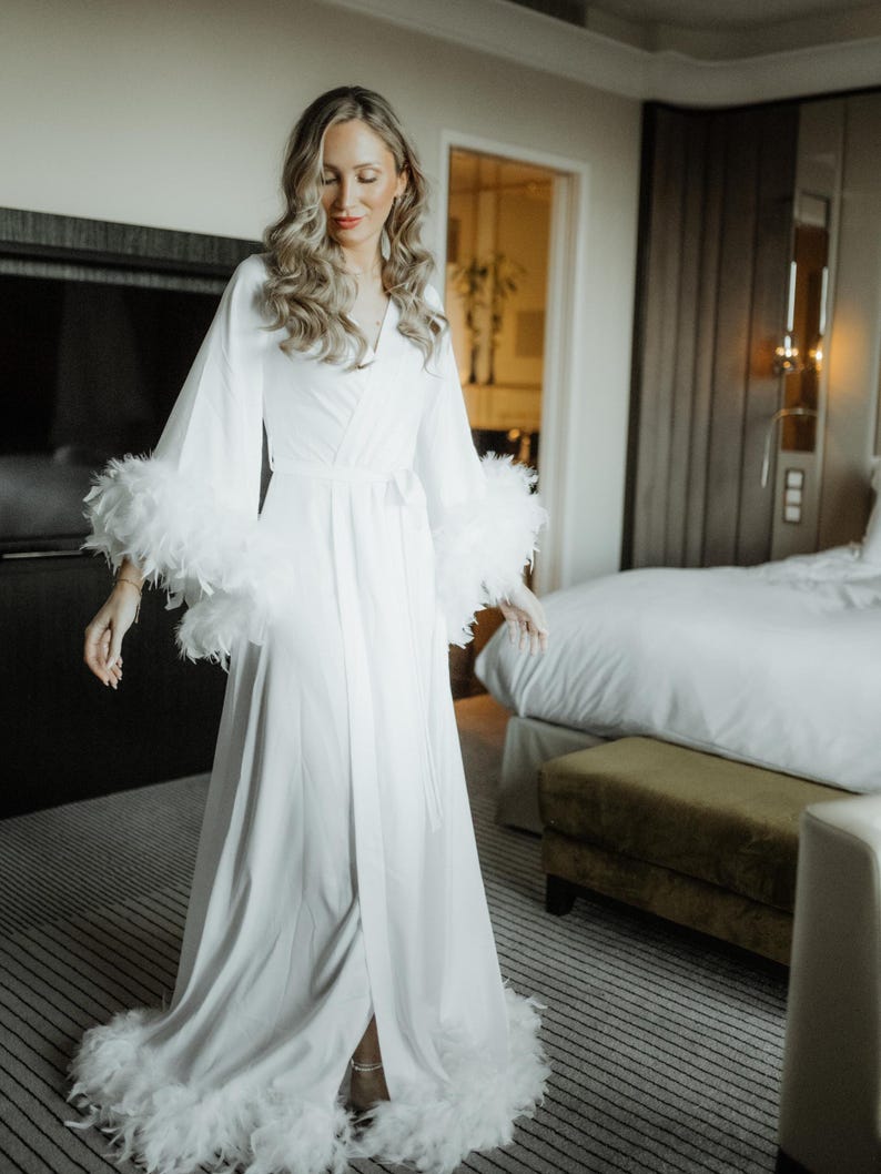 May include: A floor-length white robe featuring long, wide sleeves and a hemline trimmed with white feathers. The garment includes a wrap-style front with a matching fabric tie belt at the waist, creating a flowing silhouette with soft, draped fabric.
