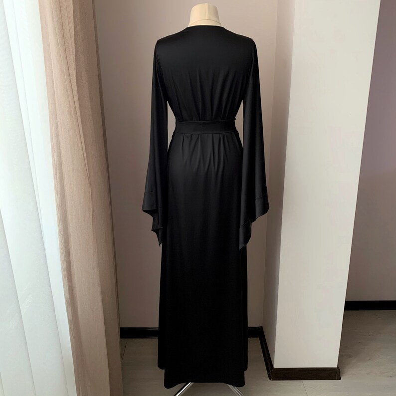 Puede incluir: Un vestido largo negro con mangas largas y acampanadas y un cintur&oacute;n a juego. El vestido se muestra en un maniqu&iacute;, mostrando su silueta fluida y su dise&ntilde;o elegante. La tela parece ser un material suave y drapeado.