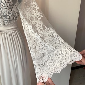 May include: White maxi dress with long sleeves featuring a delicate floral lace pattern. The sleeves are sheer and have a bell shape.