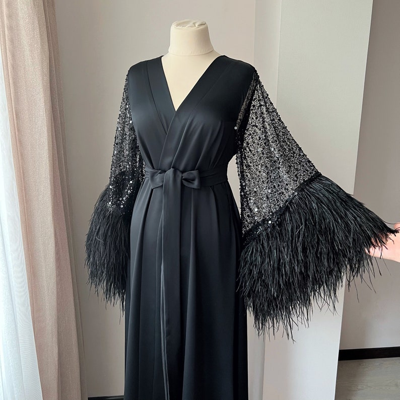K&ouml;nnte beinhalten: Eine schwarze Satin-Robe mit langen &Auml;rmeln, die mit schwarzem Federnbesatz und Pailletten verziert sind.