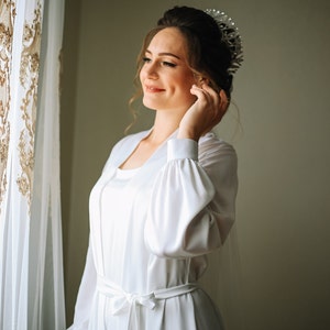 May include: A bride in a white satin robe with a tiara on her head, looking to the side and smiling.