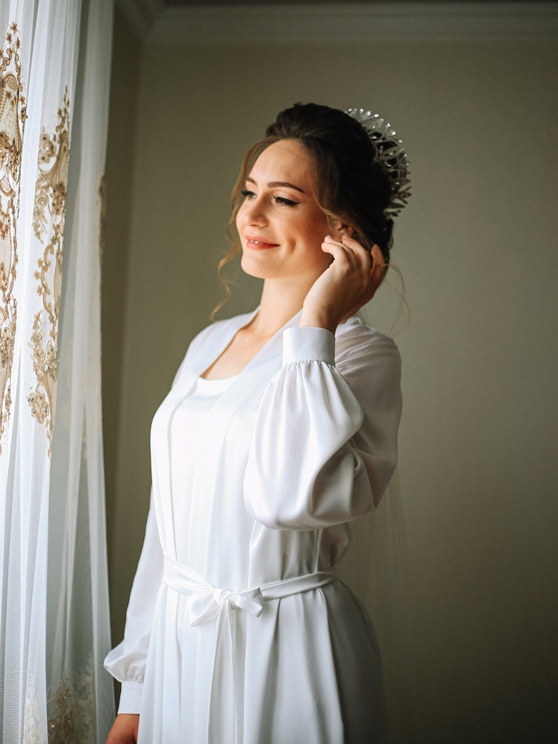 May include: A bride in a white satin robe with a tiara on her head, looking to the side and smiling.