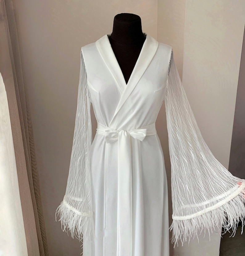 May include: A white satin robe with long sleeves and a lace overlay. The sleeves have feather trim. The robe has a sash tie at the waist.