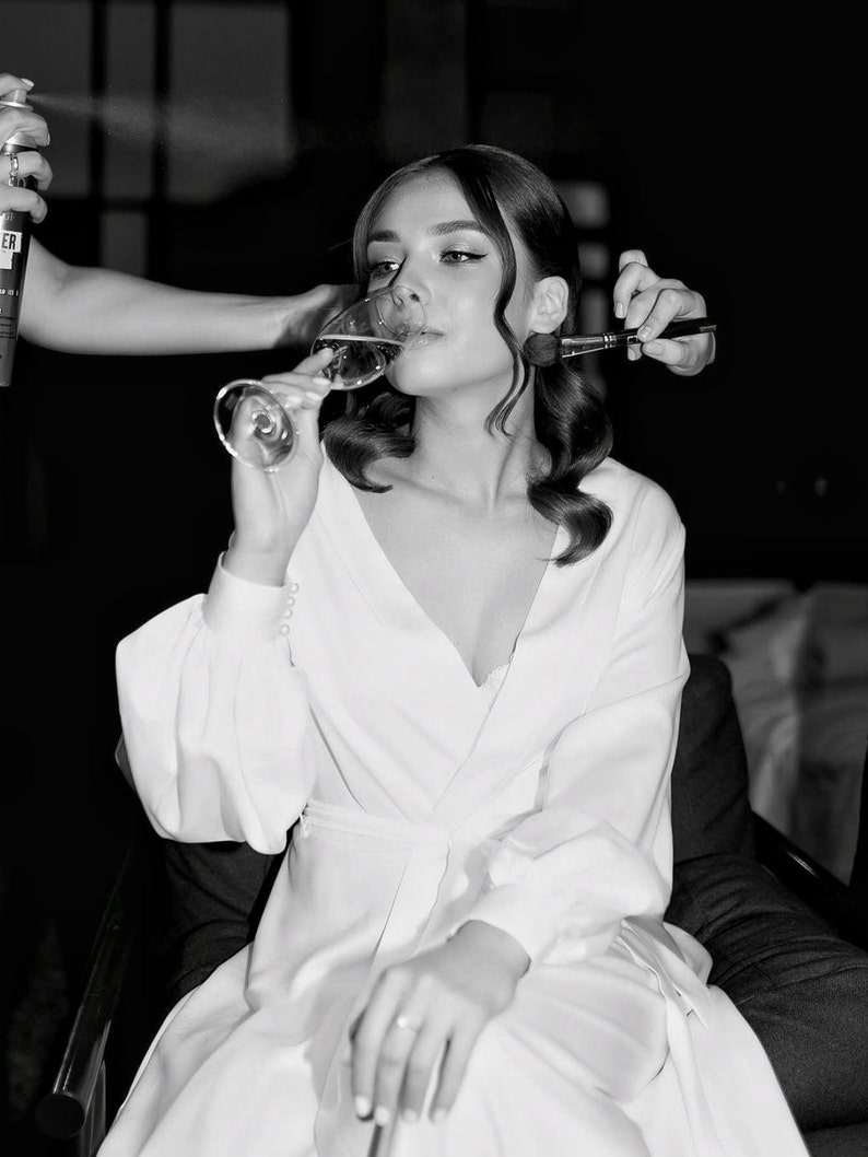 May include: A woman in a white robe sits in a chair and drinks from a champagne flute. She is having her makeup done by a makeup artist.
