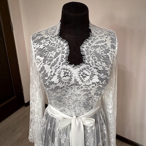 May include: A white lace robe with long sleeves and a satin ribbon belt tied in a bow. The robe is displayed on a black mannequin. The lace features a floral pattern.