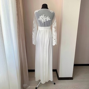 May include: White maxi dress with long lace sleeves and a floral lace design on the back. The dress has a fitted bodice and a flowing skirt.