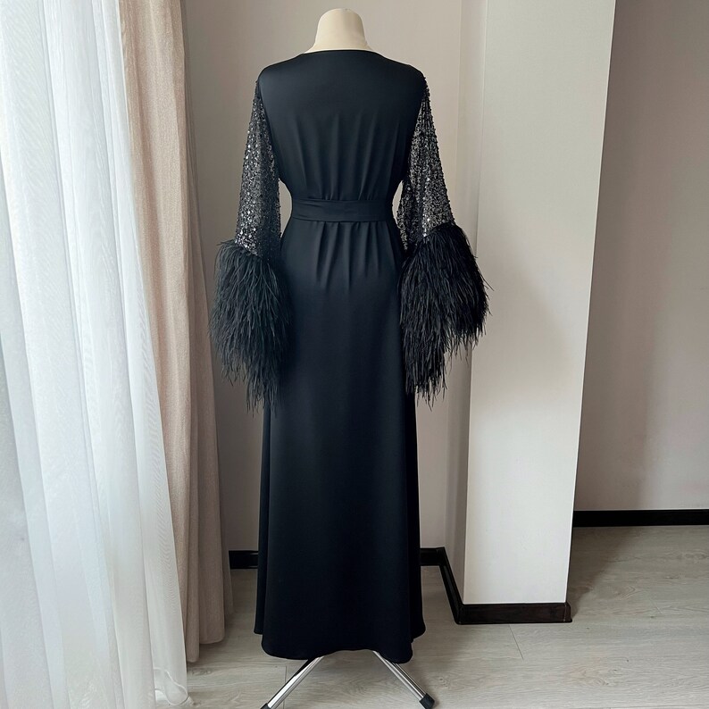 K&ouml;nnte beinhalten: Ein schwarzes Maxikleid mit langen &Auml;rmeln, die mit Federn besetzt sind und mit Pailletten verziert sind. Das Kleid hat einen Wickelstil und eine Bindeg&uuml;rtel an der Taille.