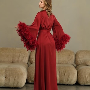 May include: A long, red satin robe with feather trim on the sleeves. The robe has a sash tie at the waist and is shown on a model.