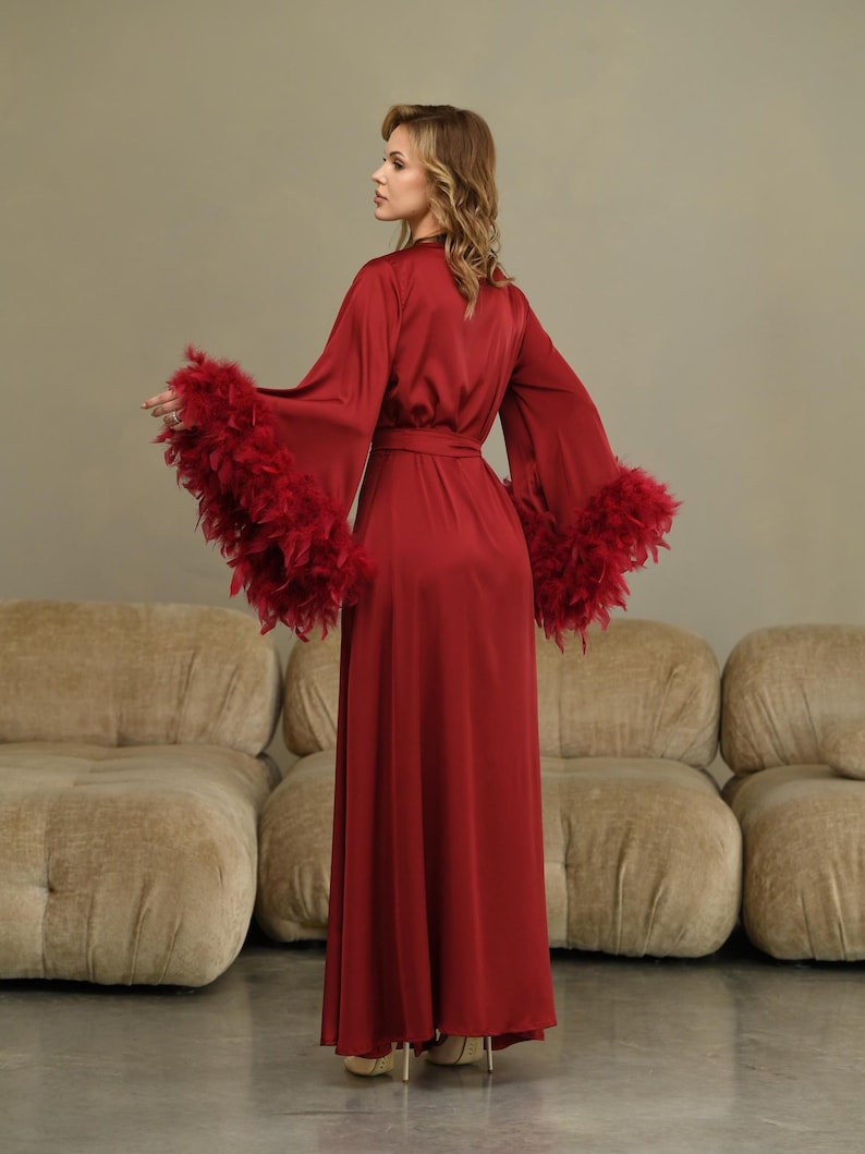 May include: A long, red satin robe with feather trim on the sleeves. The robe has a sash tie at the waist and is shown on a model.