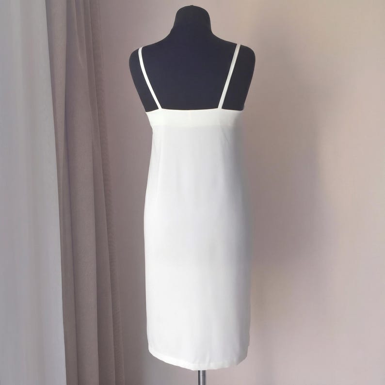 May include: A white slip dress with thin spaghetti straps and a straight neckline. The dress is displayed on a black mannequin against a neutral background. The dress appears to be made of a lightweight, flowing fabric.