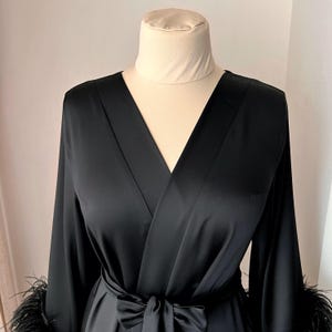 May include: A black satin robe with long sleeves and a V-neckline. The robe has a sash tie at the waist and feather trim on the cuffs.