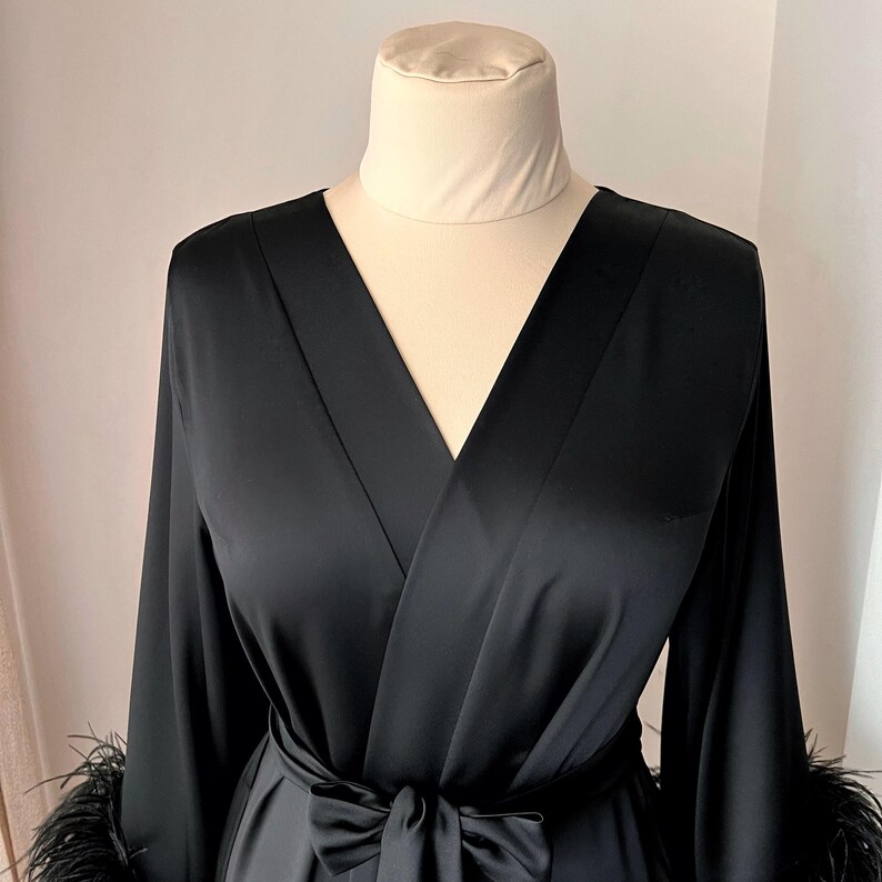 May include: A black satin robe with long sleeves and a V-neckline. The robe has a sash tie at the waist and feather trim on the cuffs.