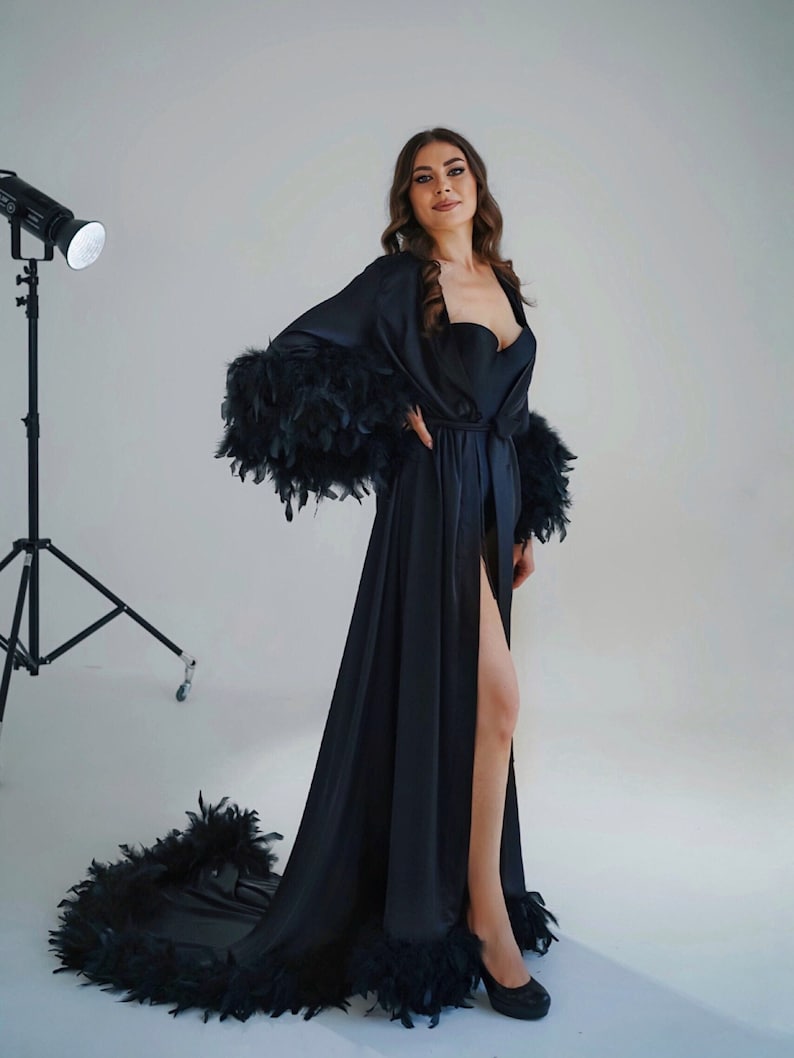 May include: A long black satin robe with a feather trim around the edges. The robe has a sash tie at the waist and a thigh-high slit.