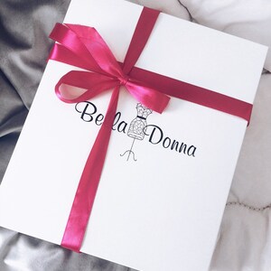 May include: A white gift box tied with a magenta ribbon and bow. The box has the words "Bella Donna" printed in black script, along with a dress form illustration. The box is on a white and gray background.