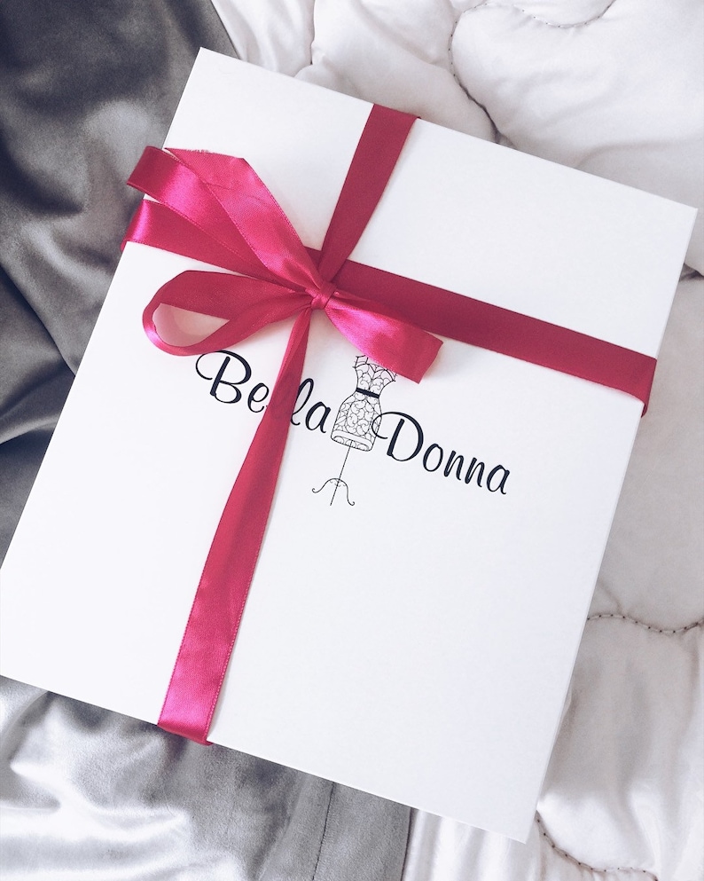 May include: A white gift box tied with a magenta ribbon and bow. The box has the words "Bella Donna" printed in black script, along with a dress form illustration. The box is on a white and gray background.
