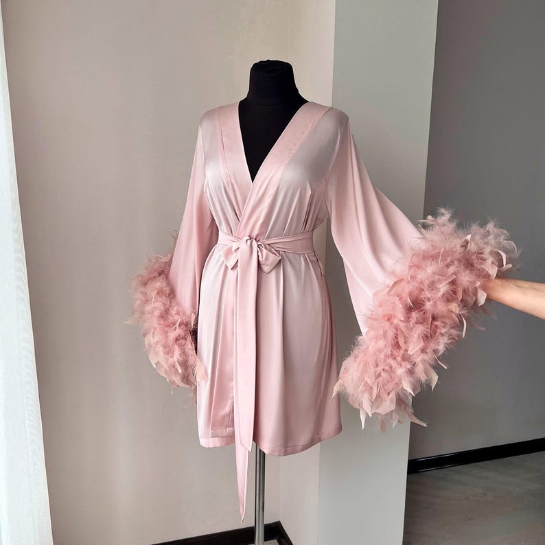 May include: A blush pink satin robe with long sleeves and a tie belt. The sleeves are trimmed with a fluffy pink feather trim.