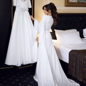 May include: A bride in a white satin robe stands in a hotel room, looking at a white wedding dress hanging on a hanger. The dress has a sweetheart neckline and a full skirt. The room has a black and gold patterned carpet and a white bed with a black headboard.