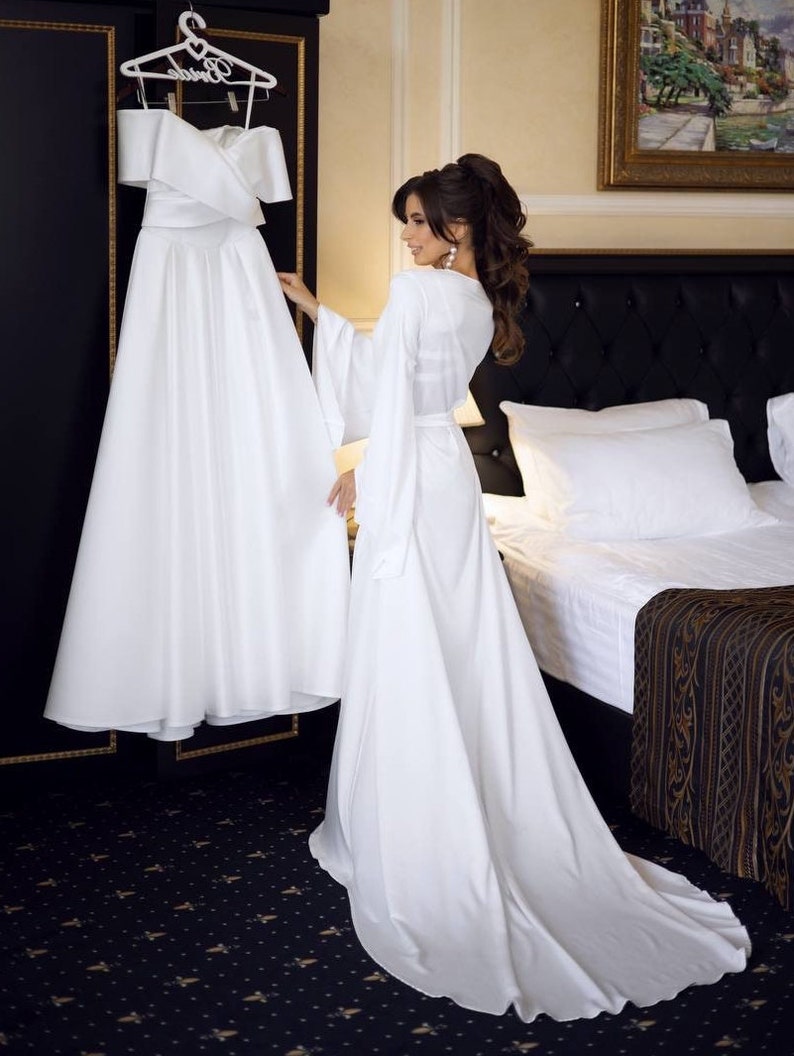May include: A bride in a white satin robe stands in a hotel room, looking at a white wedding dress hanging on a hanger. The dress has a sweetheart neckline and a full skirt. The room has a black and gold patterned carpet and a white bed with a black headboard.