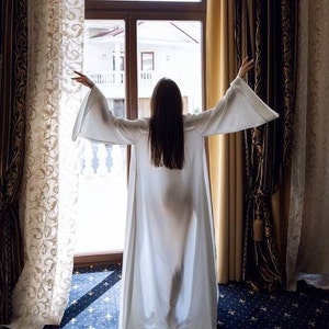 May include: A person wearing a white robe stands in front of a window with curtains, looking out at a view of a building.