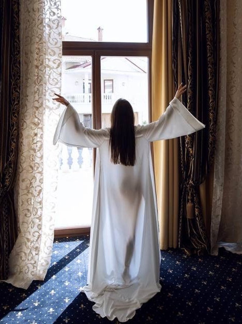 May include: A person wearing a white robe stands in front of a window with curtains, looking out at a view of a building.