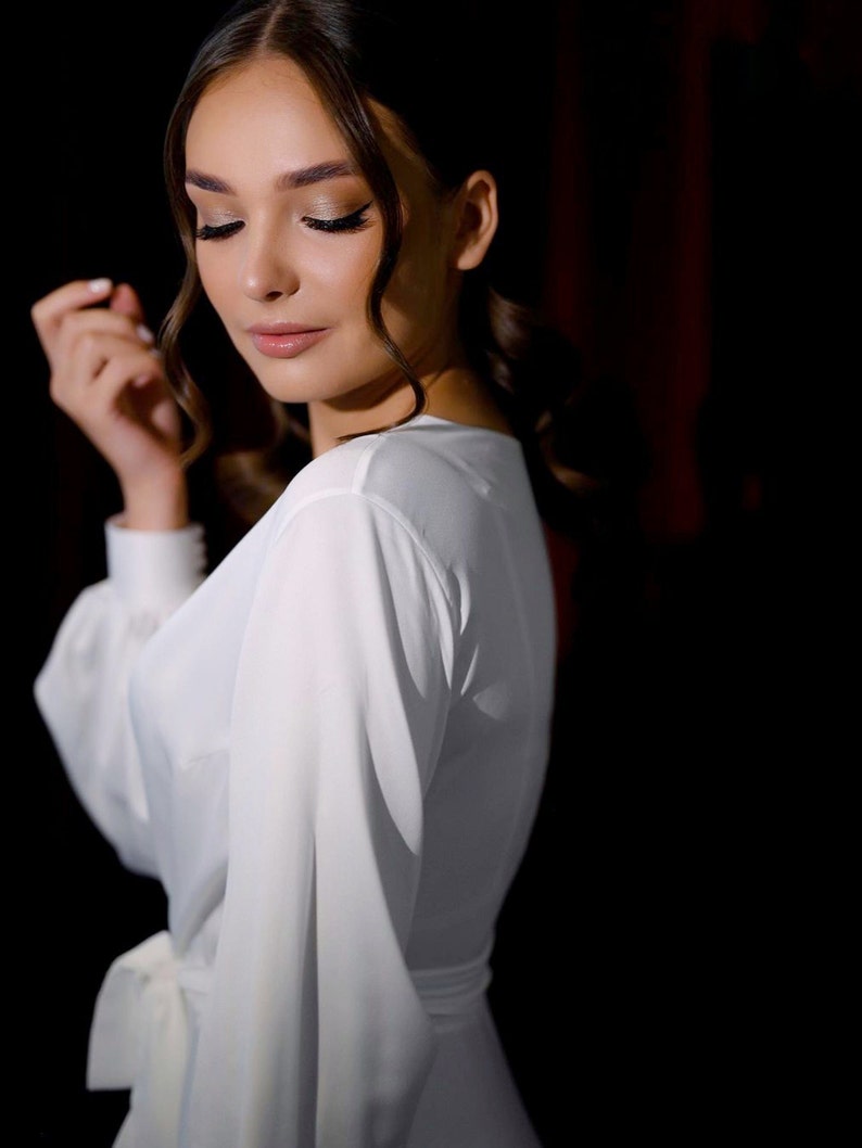 May include: A woman wearing a white, long-sleeved dress with a tie at the waist. The dress has a V-neckline and the woman is looking down.