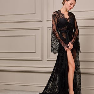 Model in a floor-length black lace robe with flared sleeves, delicate detailing, and a satin sash, standing in an elegant interior setting.