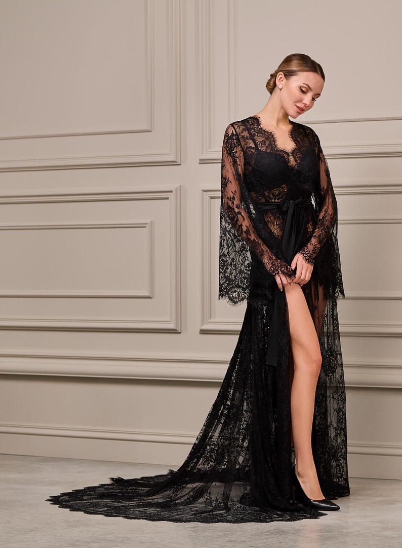 Model in a floor-length black lace robe with flared sleeves, delicate detailing, and a satin sash, standing in an elegant interior setting.