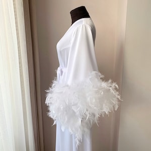 May include: A white robe featuring long, wide sleeves trimmed with thick, voluminous white feathers. The garment includes a matching white fabric tie belt at the waist and is displayed on a mannequin with a dark brown neck covering.