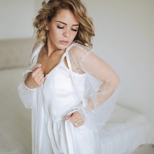 K&ouml;nnte beinhalten: Ein wei&szlig;er Satin-Shorts-Set mit einer durchsichtigen, mit Perlen besetzten Robe. Die Robe hat lange &Auml;rmel und ein zartes, feminines Design.