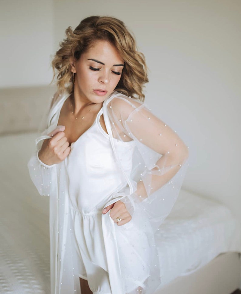 K&ouml;nnte beinhalten: Ein wei&szlig;er Satin-Shorts-Set mit einer durchsichtigen, mit Perlen besetzten Robe. Die Robe hat lange &Auml;rmel und ein zartes, feminines Design.