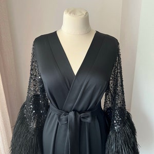 K&ouml;nnte beinhalten: Eine schwarze Satin-Robe mit langen &Auml;rmeln, die mit schwarzen Pailletten und Federn besetzt sind. Die Robe hat einen V-Ausschnitt und eine Bindeg&uuml;rtel an der Taille.