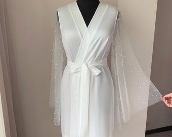 May include: White satin robe with a V-neck and a matching tie belt. The sheer sleeves are adorned with small, scattered pearls. The robe is displayed on a black mannequin head.
