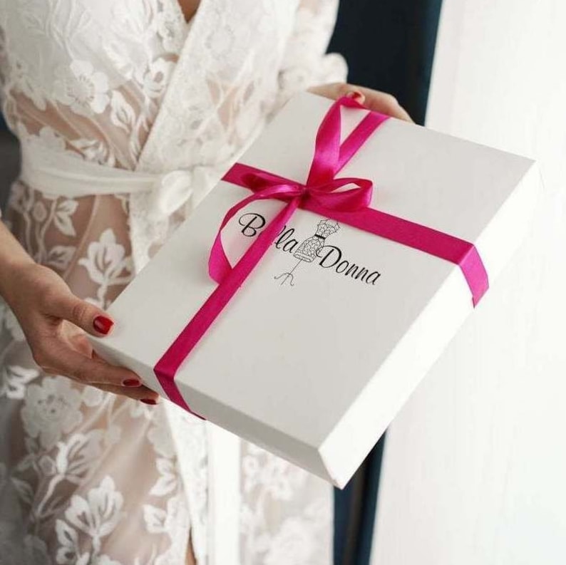 May include: A white gift box with a pink ribbon and bow. The box has a logo that says "Bella Donna" with a stylized image of a dress form.