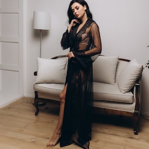 May include: A woman wearing a black sheer robe with lace detailing. The robe is open in the front, revealing a black lace bra underneath. The woman is standing in front of a white couch with pillows.