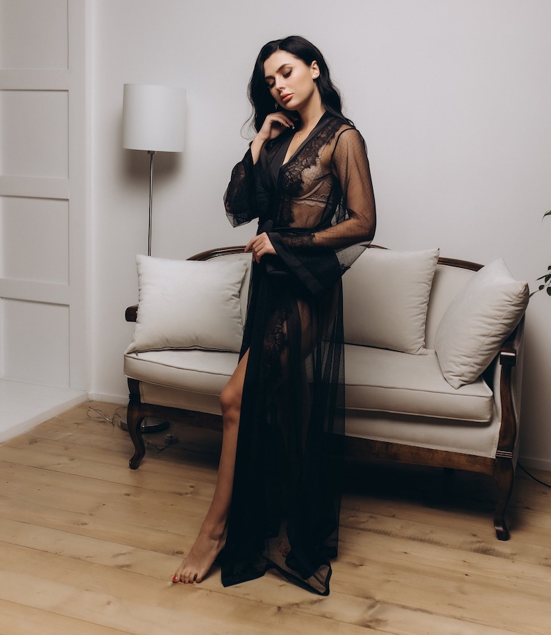 May include: A woman wearing a black sheer robe with lace detailing. The robe is open in the front, revealing a black lace bra underneath. The woman is standing in front of a white couch with pillows.