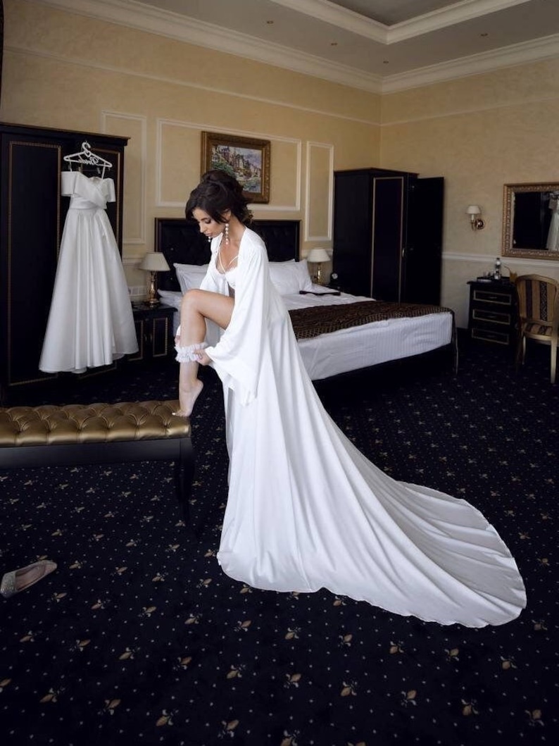 May include: A bride in a white satin robe with a long train, standing in a hotel room, putting on a garter.
