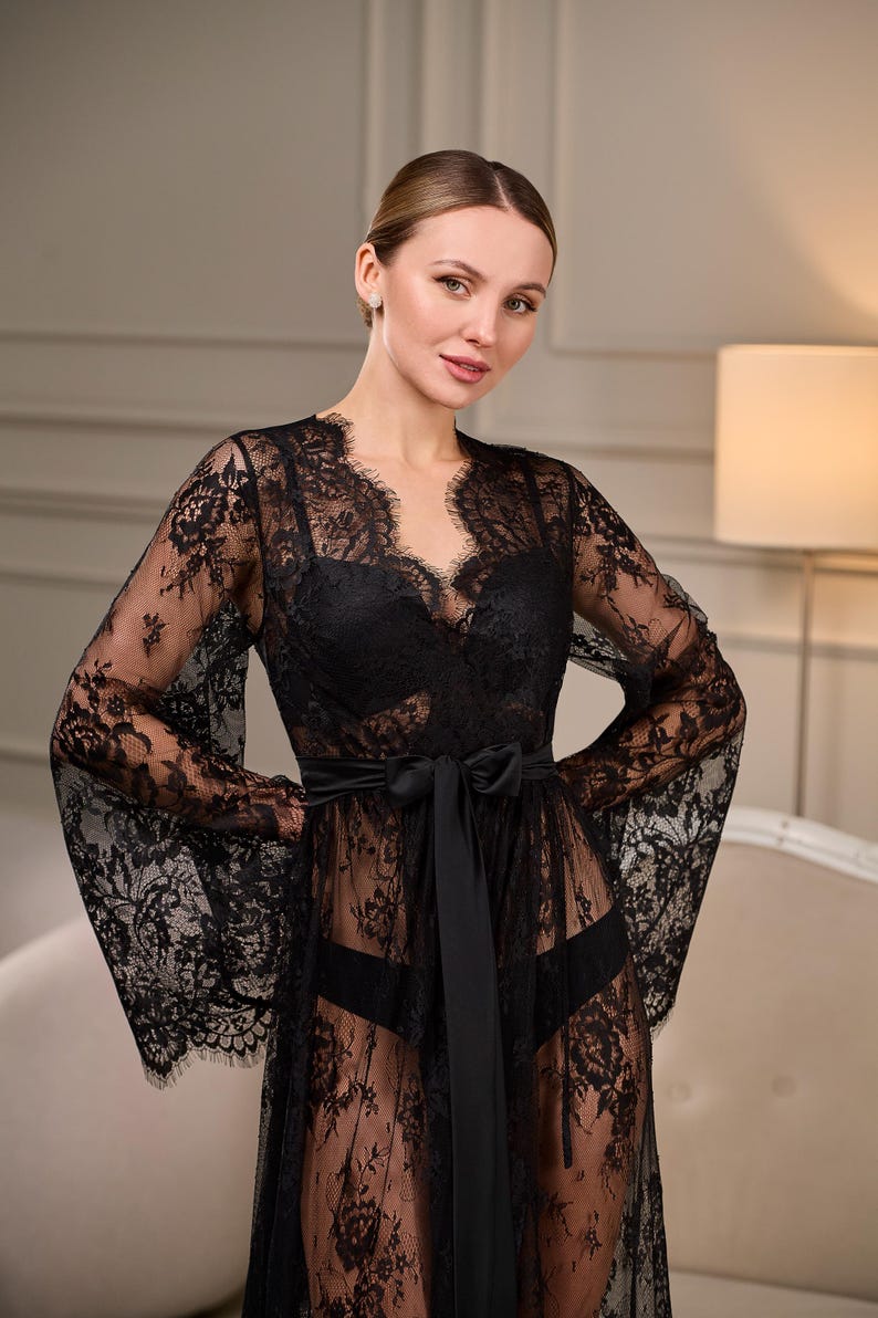 Elegant black lace robe with a dramatic train, sheer floral embroidery, and flowing bell sleeves. A satin belt cinches the waist, creating a refined silhouette.