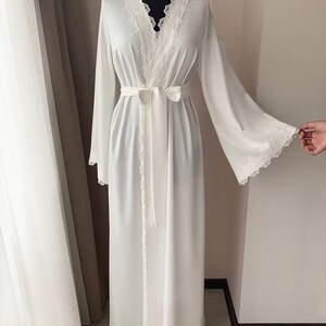 Peut inclure: Une robe blanche &agrave; manches longues avec une bordure en dentelle autour du col, des poignets et de la ceinture. La robe a un col en V et une ceinture qui se noue &agrave; la taille.